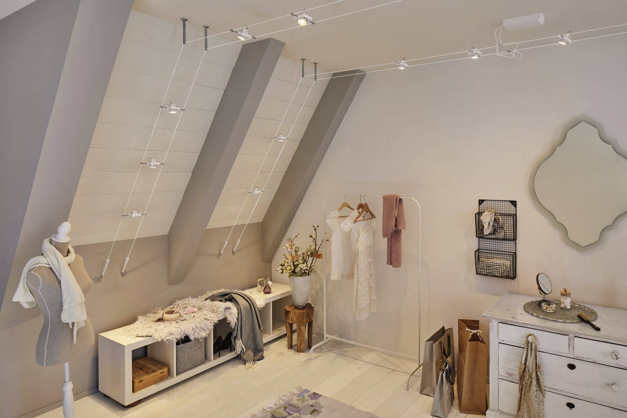 A cosy dressing room under a pitched roof, with clothing on stands, a mirror, shelves and decorative flowers.