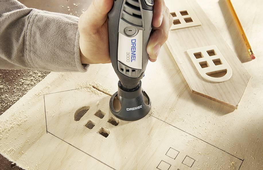 A person is using a Dremel tool to engrave window shapes into a wooden board. Sawdust is visible.