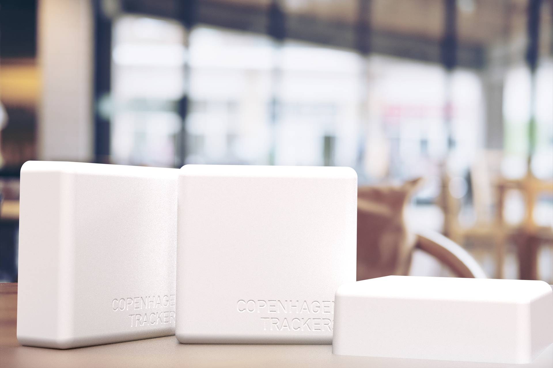 Three white, square devices on a table in a café. Inscription reads 'Copenhagen Tracker'. Background is blurred.