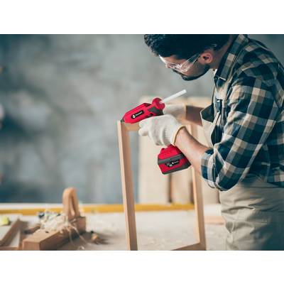 A man wearing safety glasses and gloves uses a red hot glue gun to assemble a wooden frame piece.
