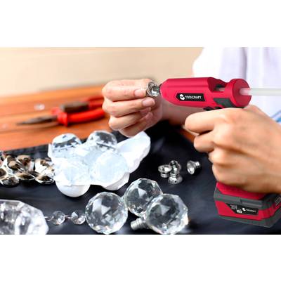 A person is using a hot glue gun to attach crystal balls and spoons to a black fabric. Pliers are lying in the background.