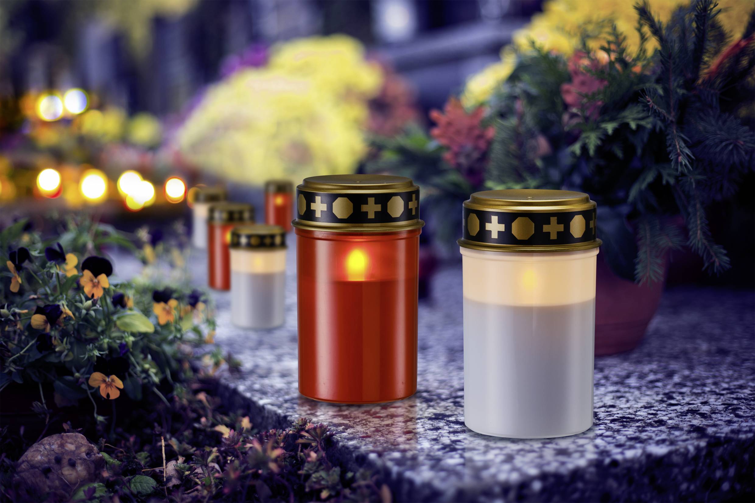 Grave candles glow amidst colourful flowers in a cemetery. They stand in a row, creating a serene and contemplative atmosphere.