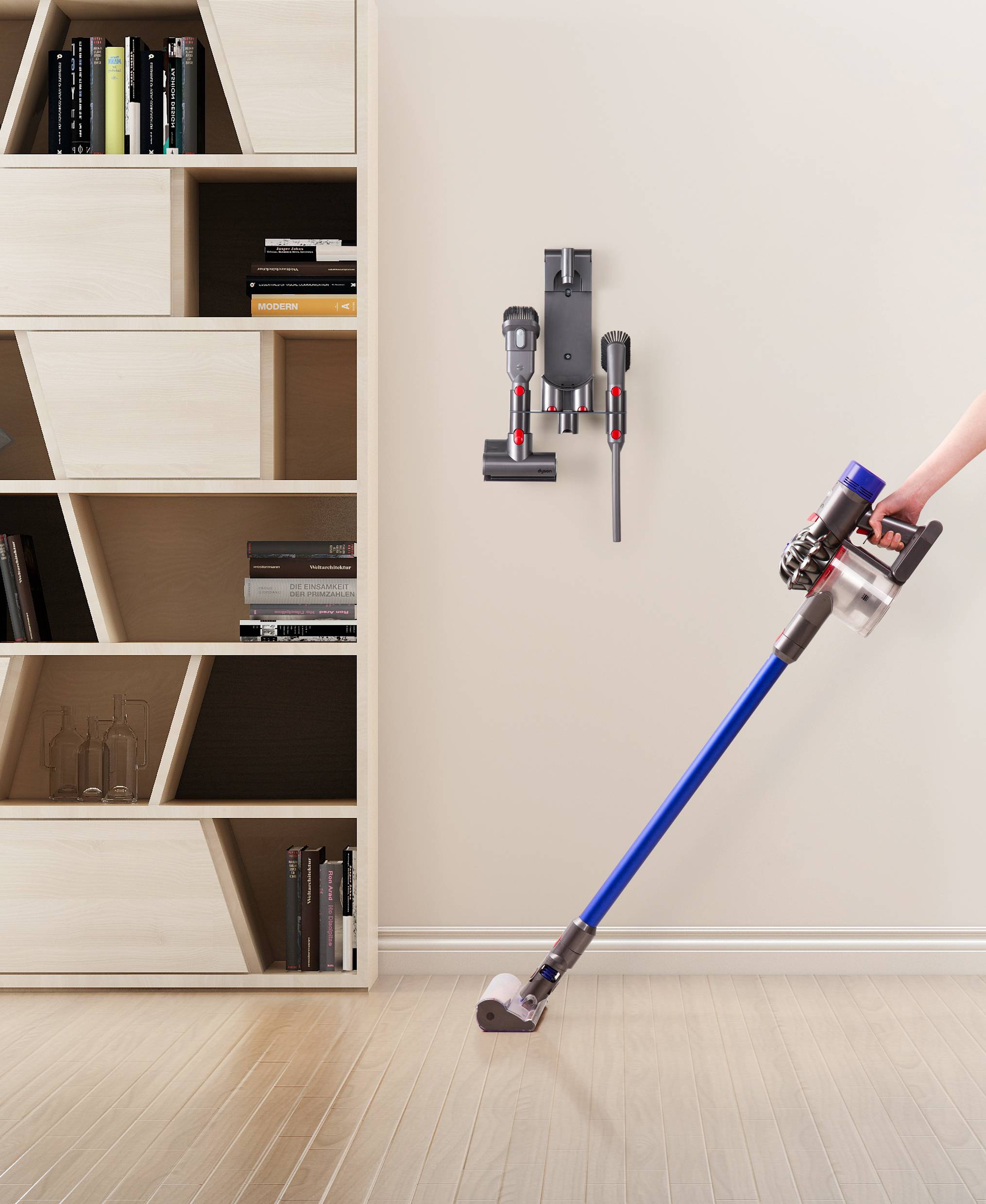 A handheld vacuum cleaner is being used by a person on a wooden floor. A charging station with accessories hangs on the wall next to a shelf.