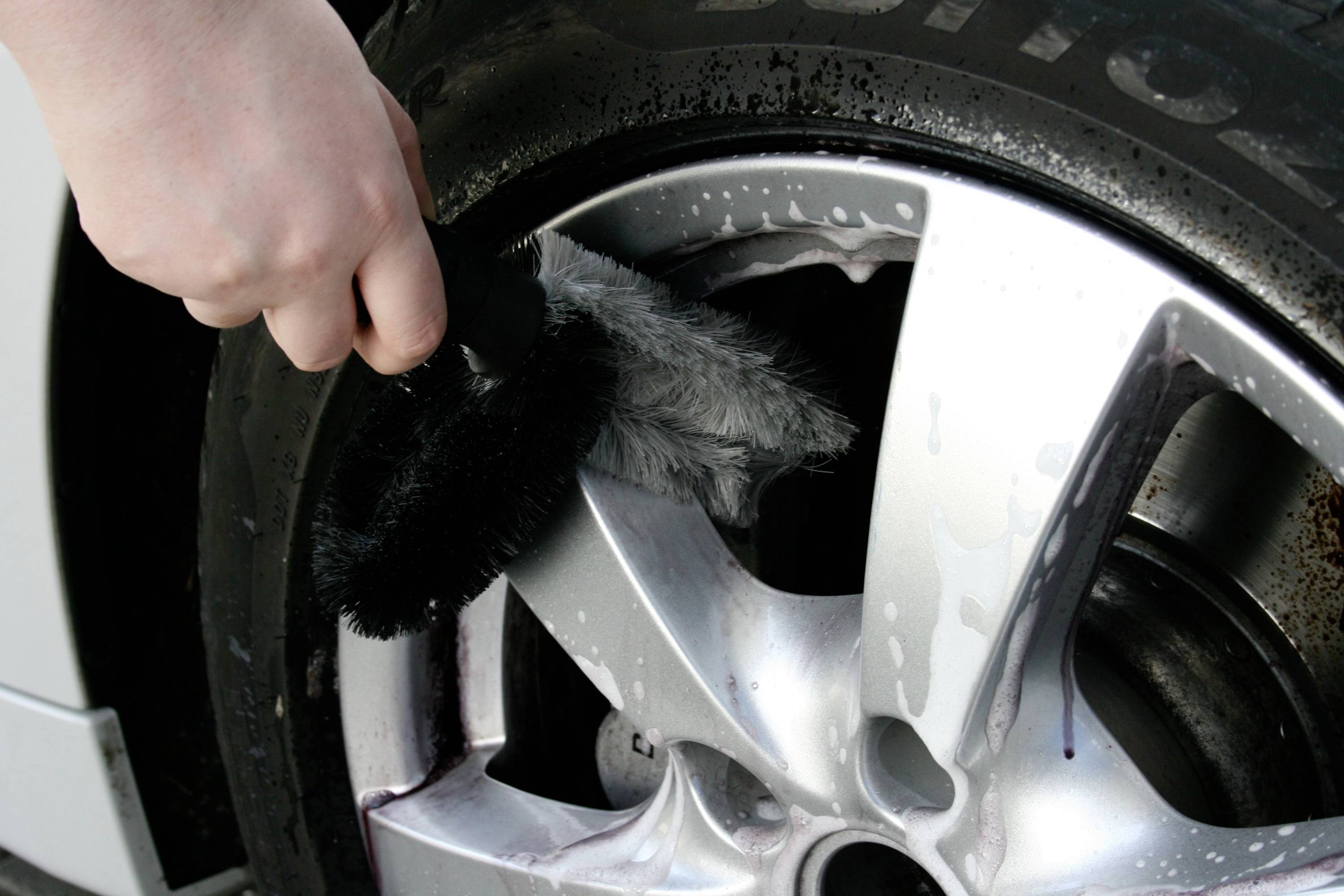 A hand is cleaning a car tyre wheel with a brush. Soap bubbles are visible, the car is being washed.