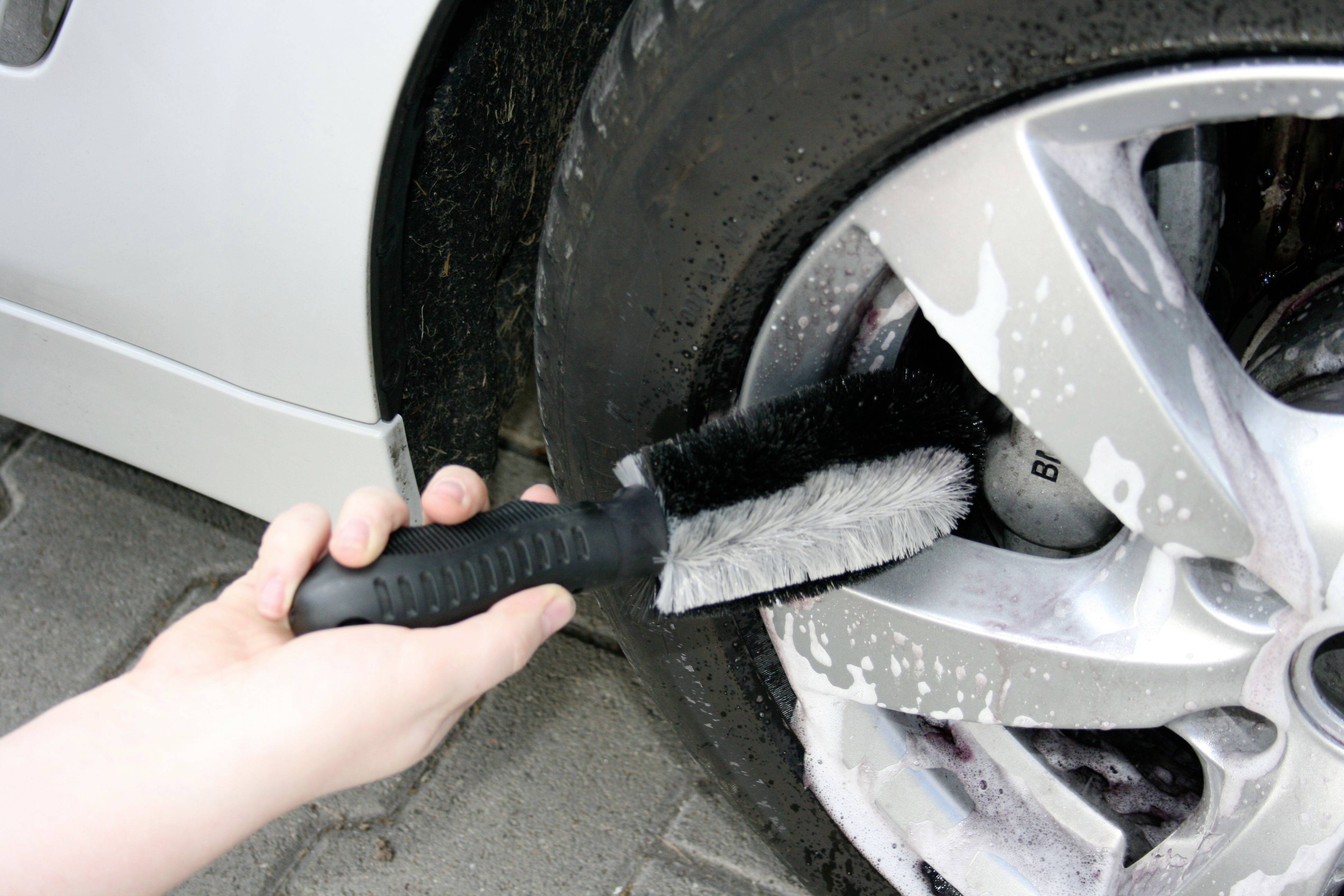 A person is cleaning the alloy wheels of a car with a brush. The wheel is covered in foam. The car is parked on a paved surface.