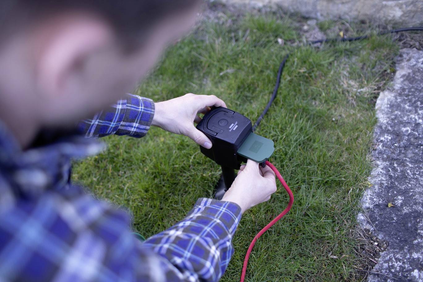 A person is installing a power socket outdoors on a grassy area. A red cable is connected.