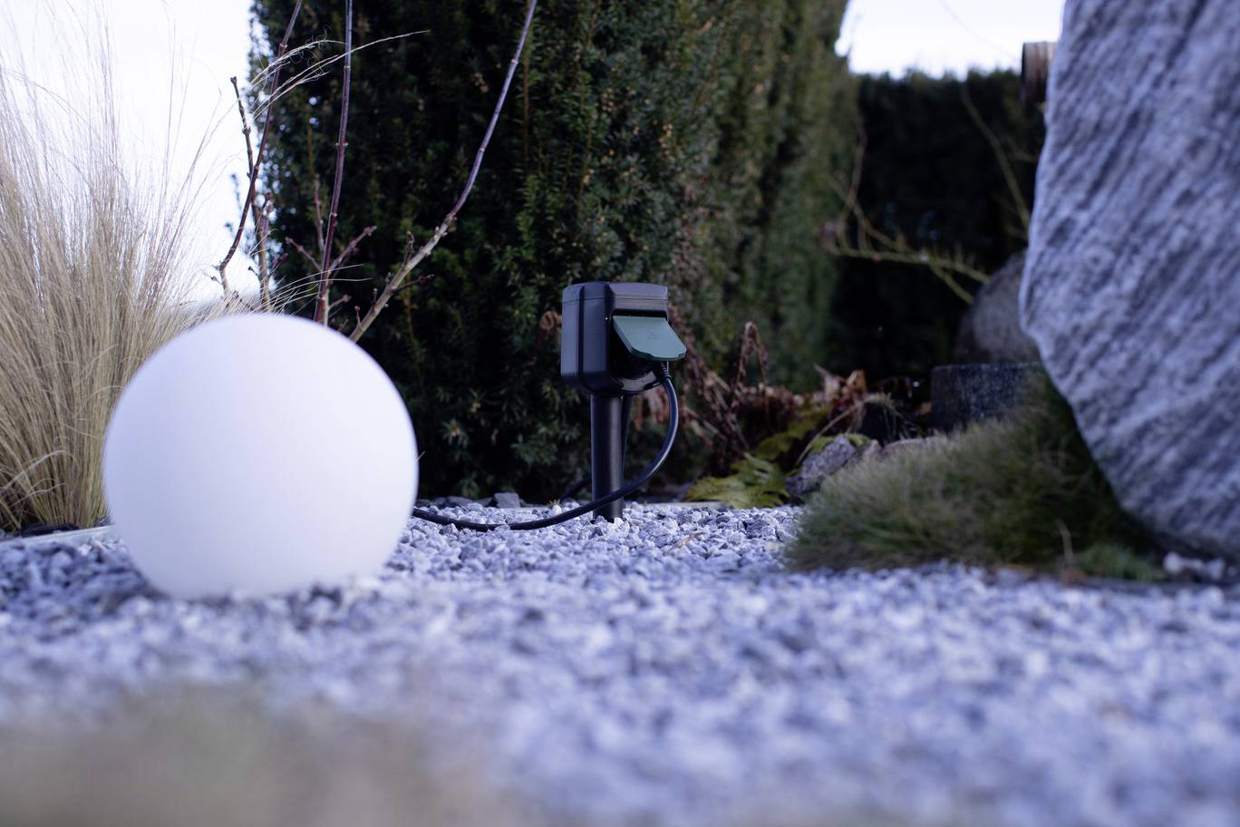 A white spherical lamp on gravel beside a power socket in the garden, surrounded by bushes and a large stone.