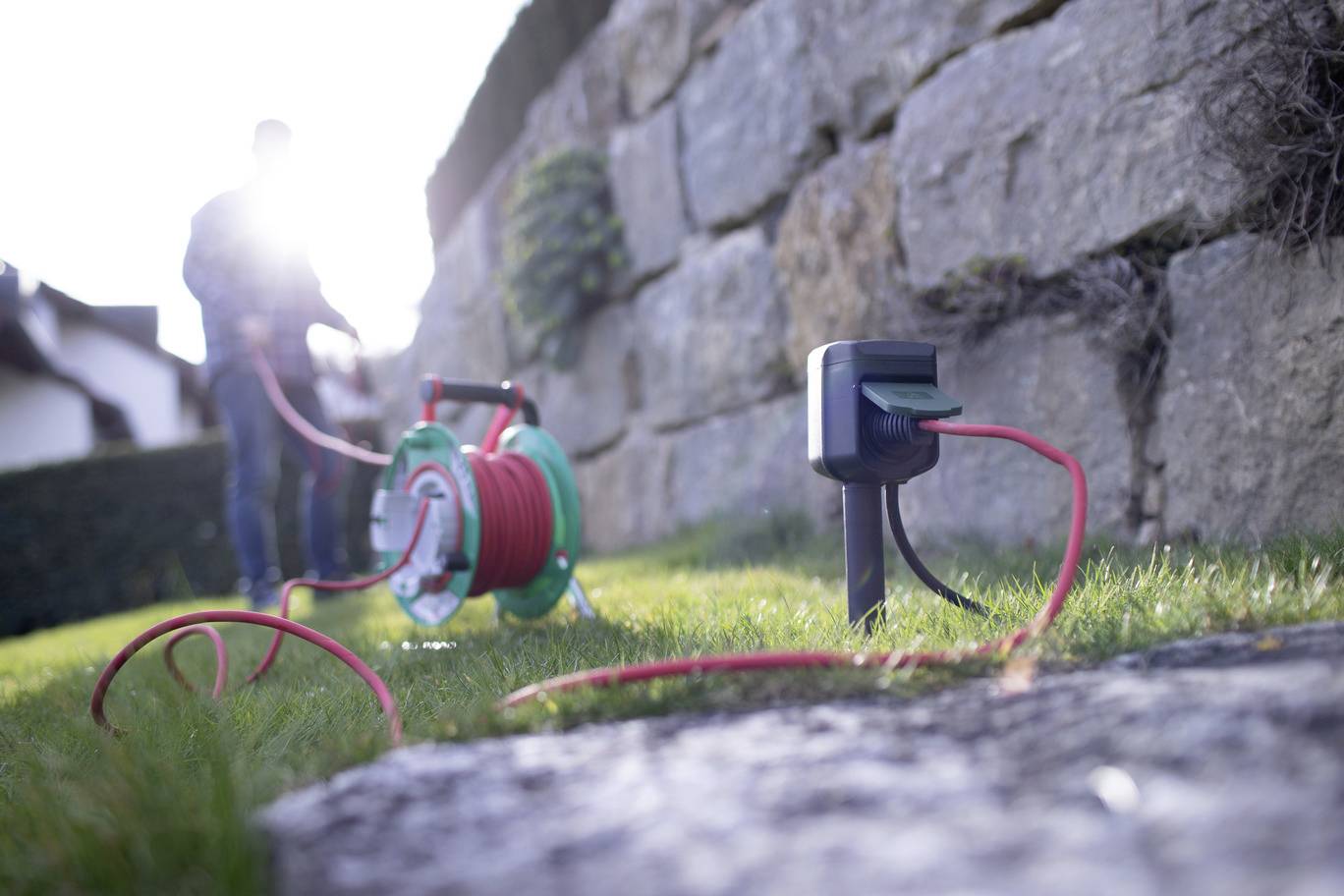A cable reel and a plug socket adapter are lying in the grass next to a stone wall. A blurred person in the background is holding a cable.