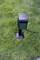 A black socket stands on green grass. A cable leads away from the socket. It is designed for outdoor use.