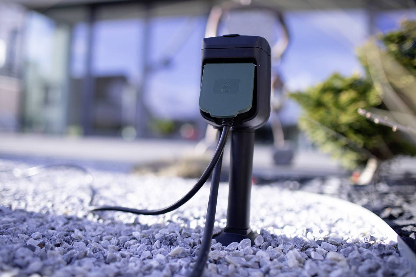 Outdoor charging station with connected cable on gravel ground, with blurred background showing buildings and plants.