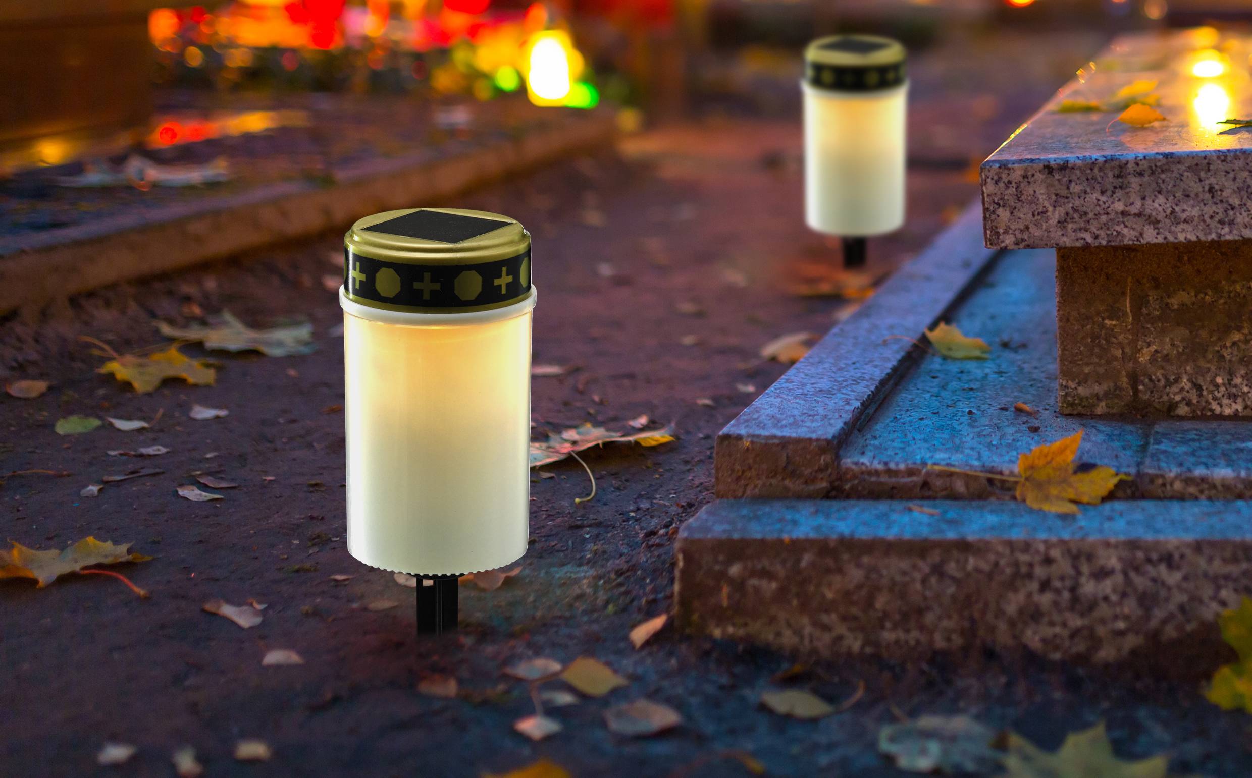 A glowing memorial candle at dusk on a cemetery, surrounded by autumn leaves. Additional memorial lights in the background.