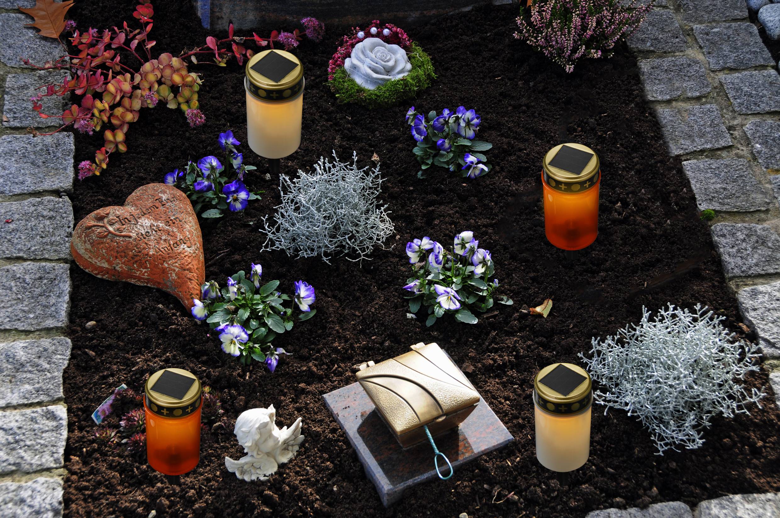 A grave with flowers and decorations, including two memorial candles, a stone heart, and an angel figurine, set on earth and framed by stones.