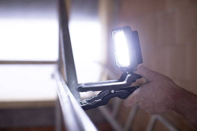 A hand holds a glowing LED work lamp with a clamp attached to a metal piece. The environment appears industrial.