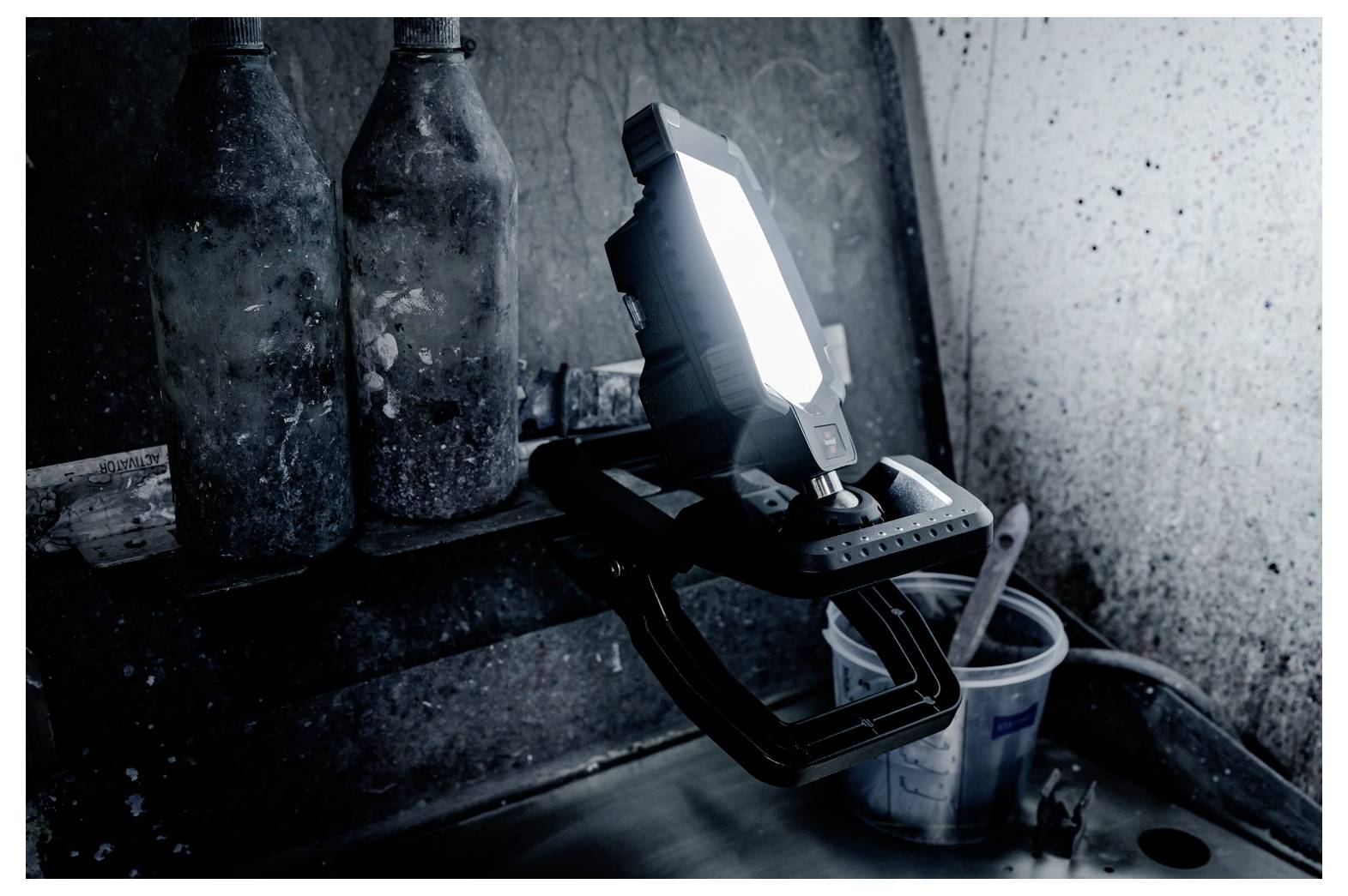 Portable work light on a stand shining in a dark, dusty industrial corner beside a bucket and hose.