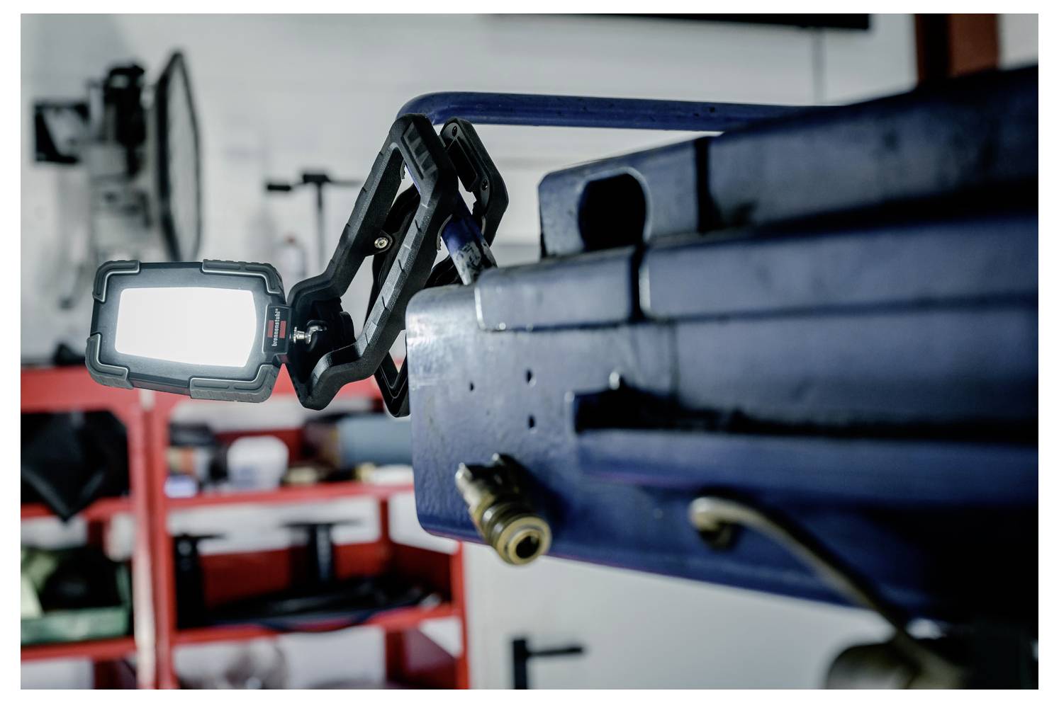 Close-up of a mounted LED work light on a blue machine arm in a workshop, with red shelving blurred in the background.
