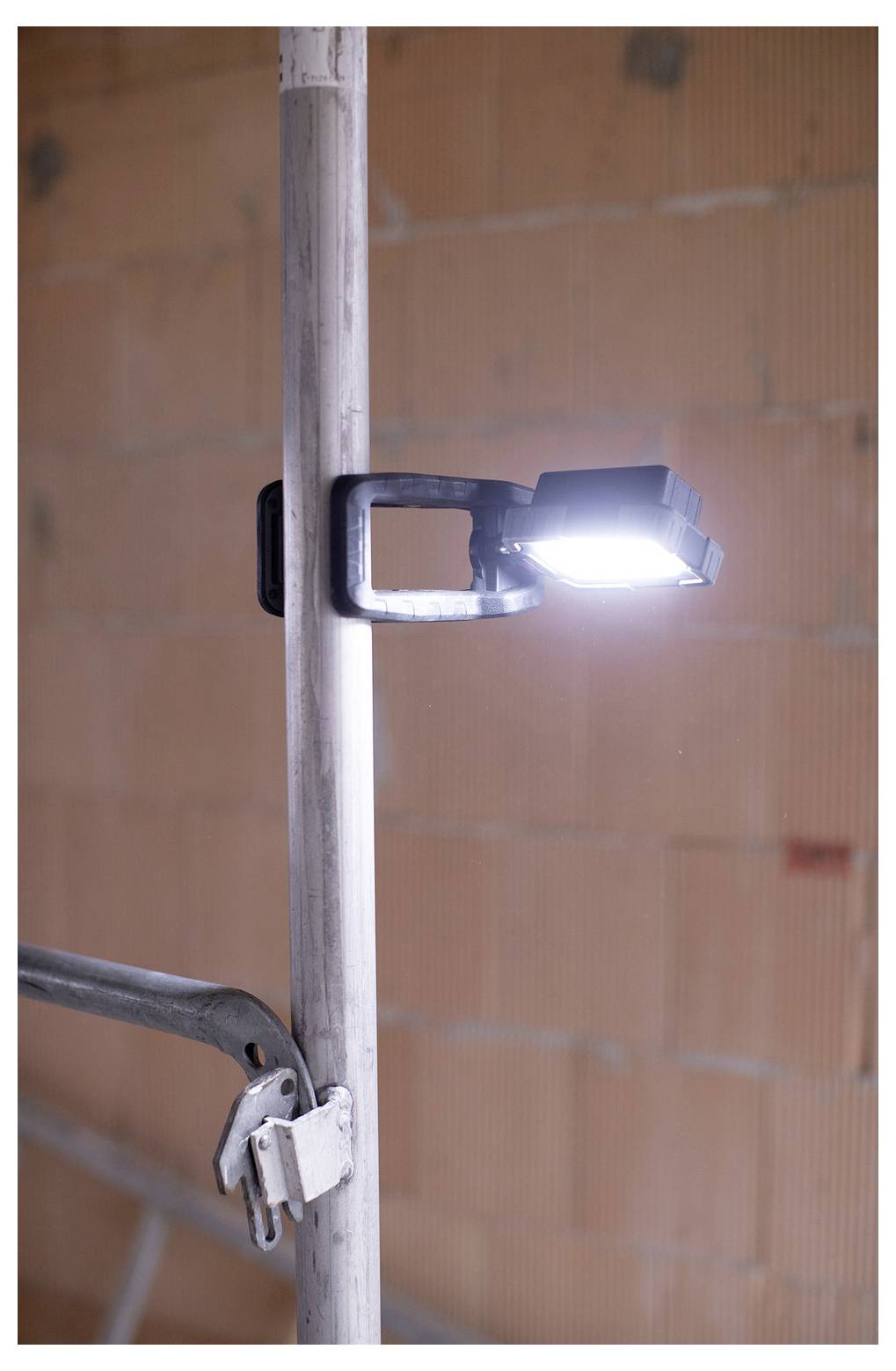 LED work light clamped to a vertical metal pole in an unfinished brick interior, with a second clamp visible below.