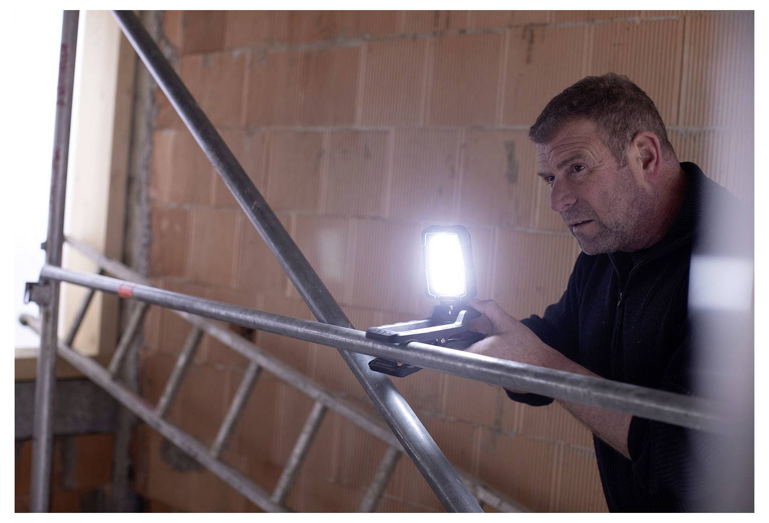 Man using a lit handheld tool or camera while inspecting scaffolding inside a brick building under construction.