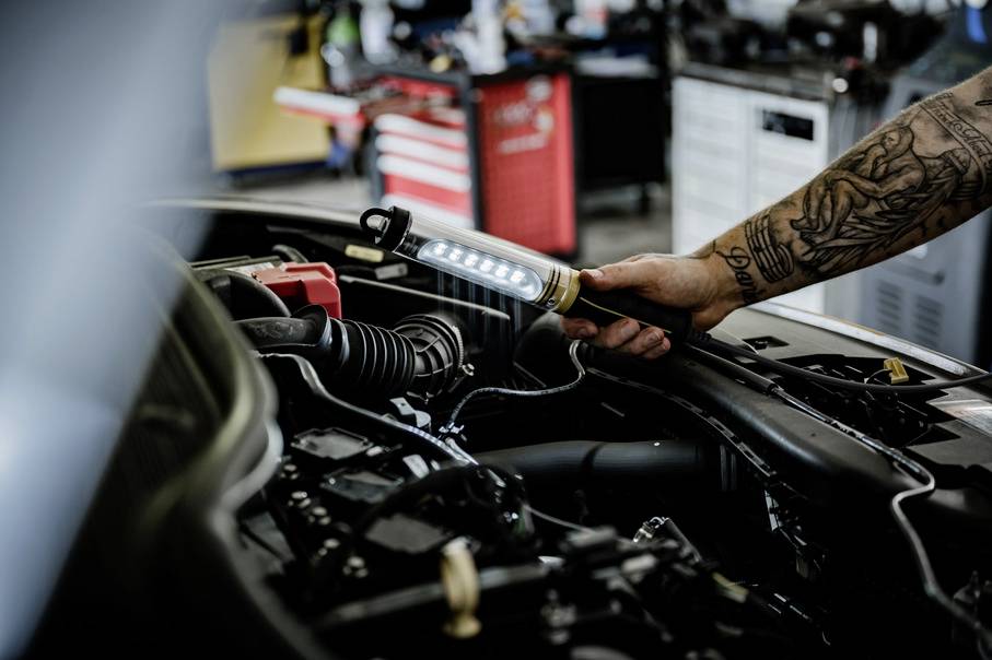 A person is illuminating the engine bay of a car in a workshop using a portable LED lamp.