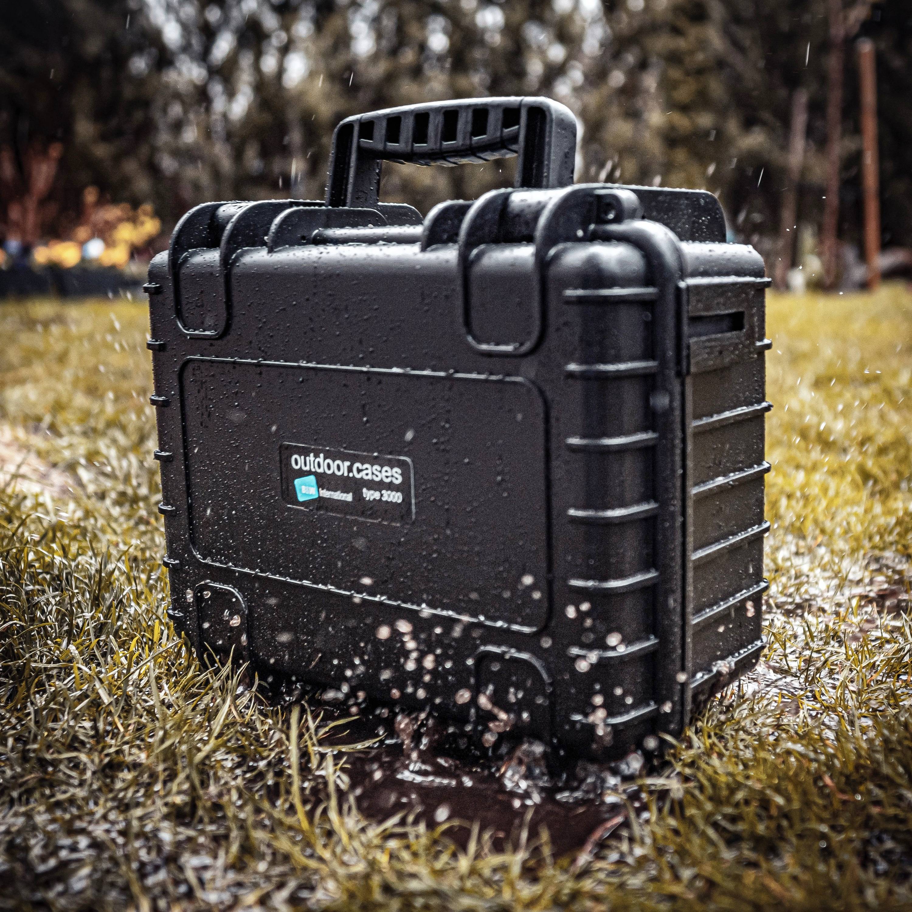 A robust, black protective case sits on damp grass, surrounded by a blurred background of trees. The case is wet with water droplets.