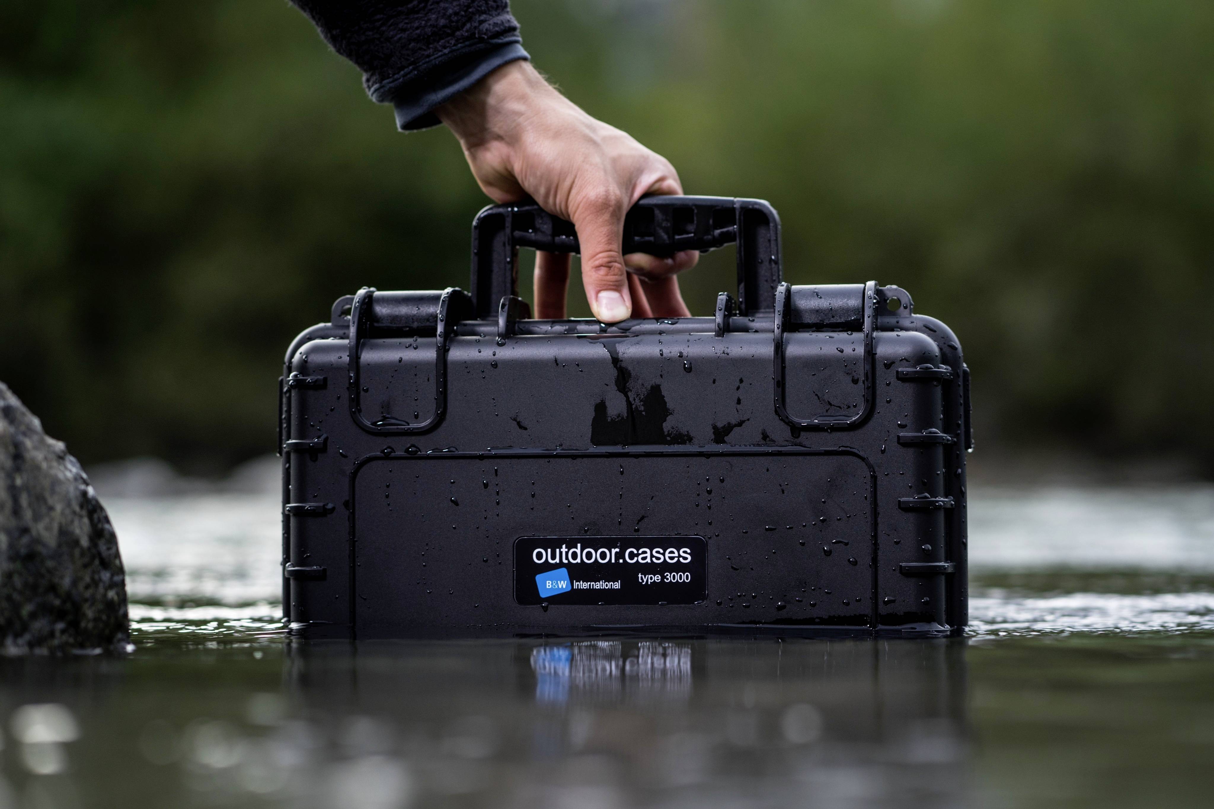 A hand is holding a black, waterproof case with the inscription 'outdoor.cases' over a calm body of water.