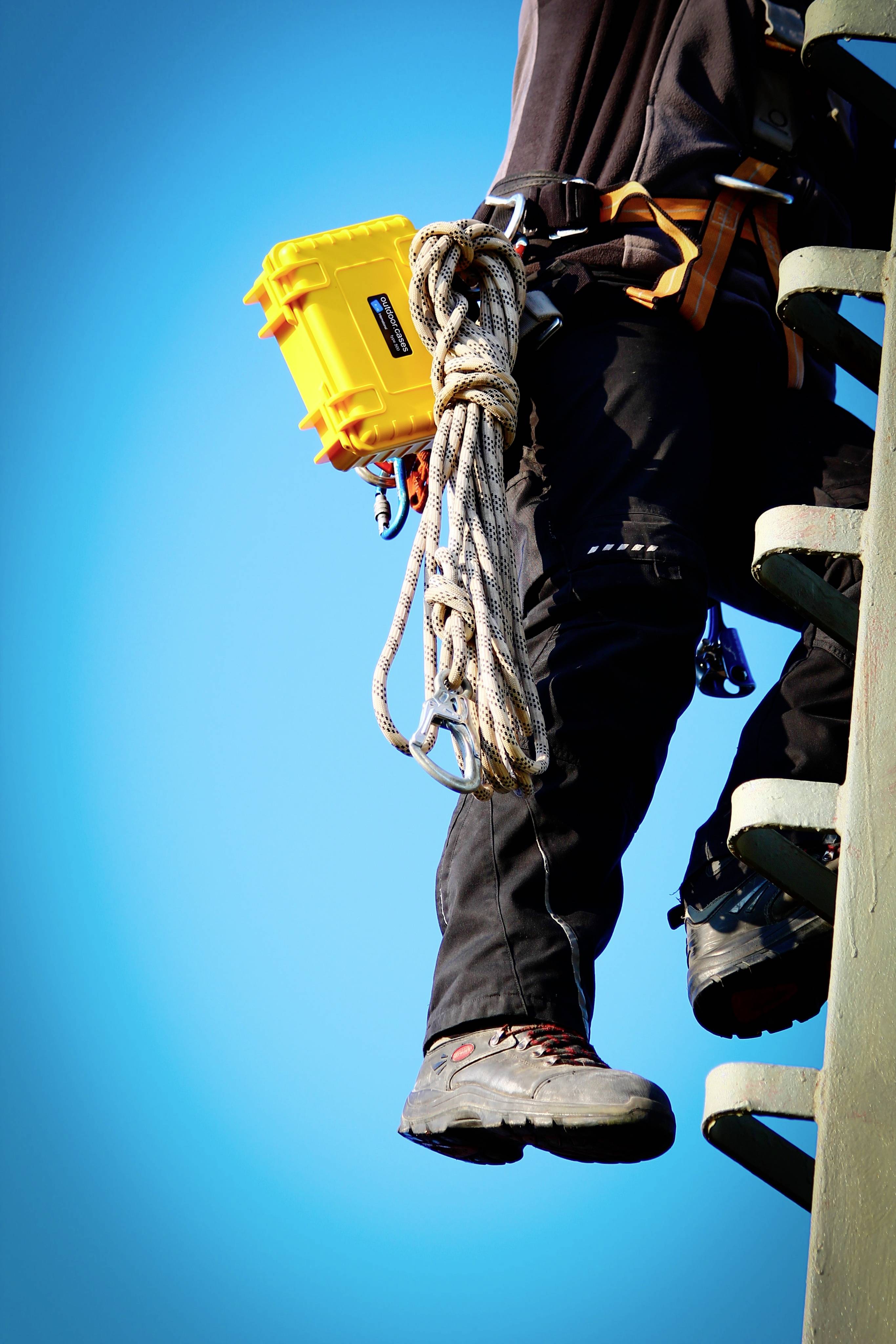 A person is climbing a ladder, wearing safety equipment and holding a yellow case, against a clear blue sky.
