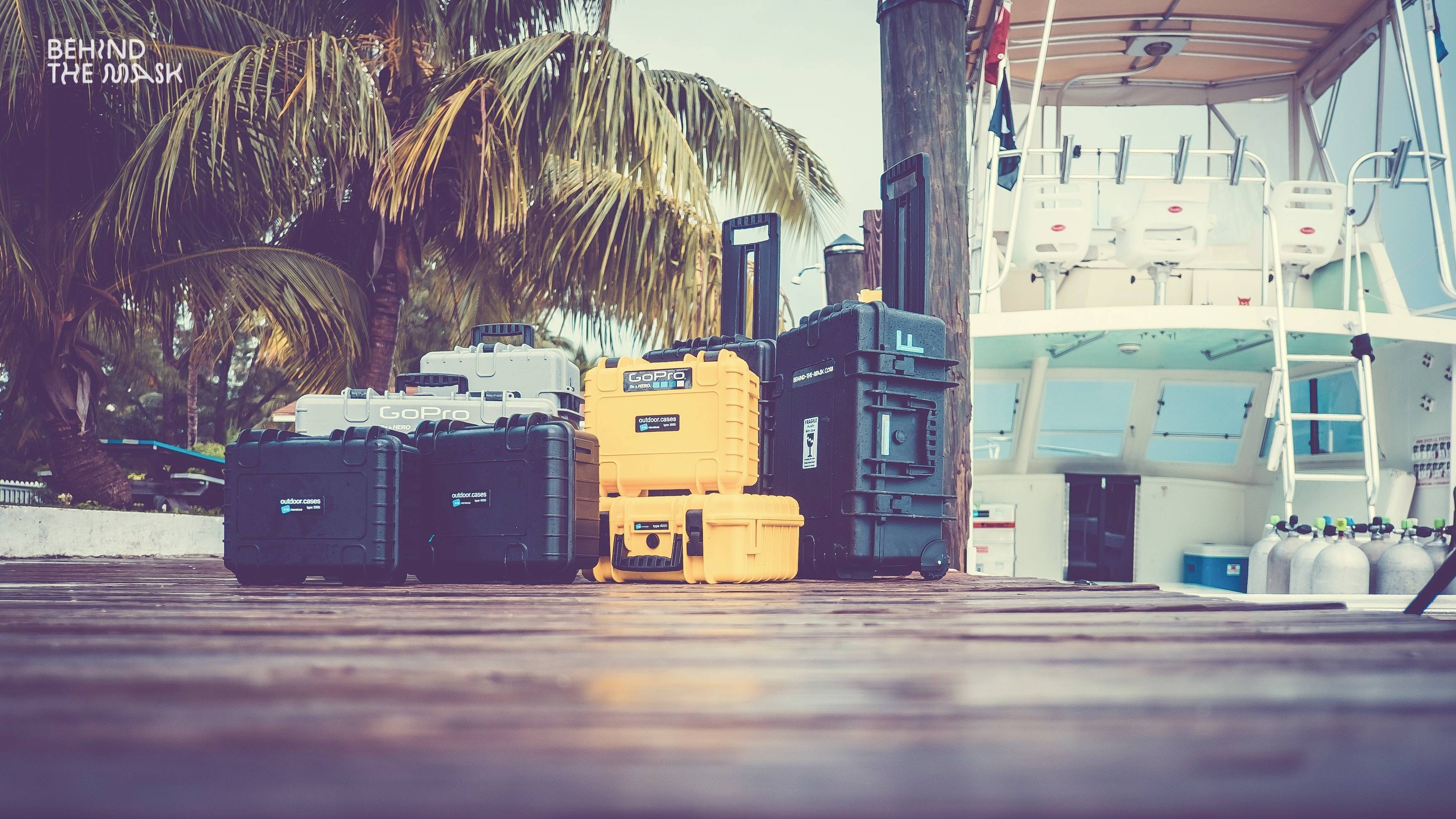 A row of equipment cases stands on a wooden jetty beside a boat in the harbour. Palms are visible in the background.
