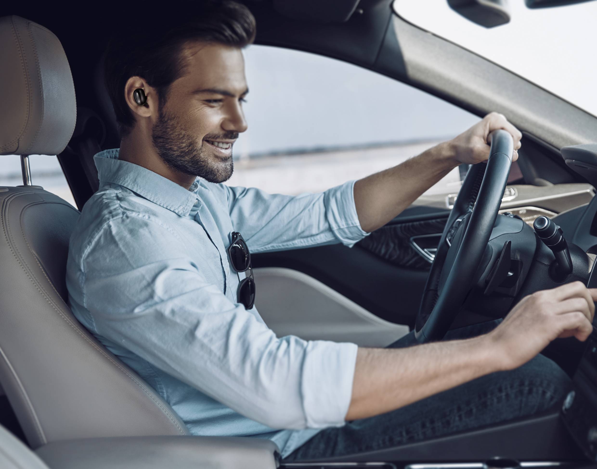 A man is driving a car with a smile, holding the steering wheel with one hand and wearing an earphone.