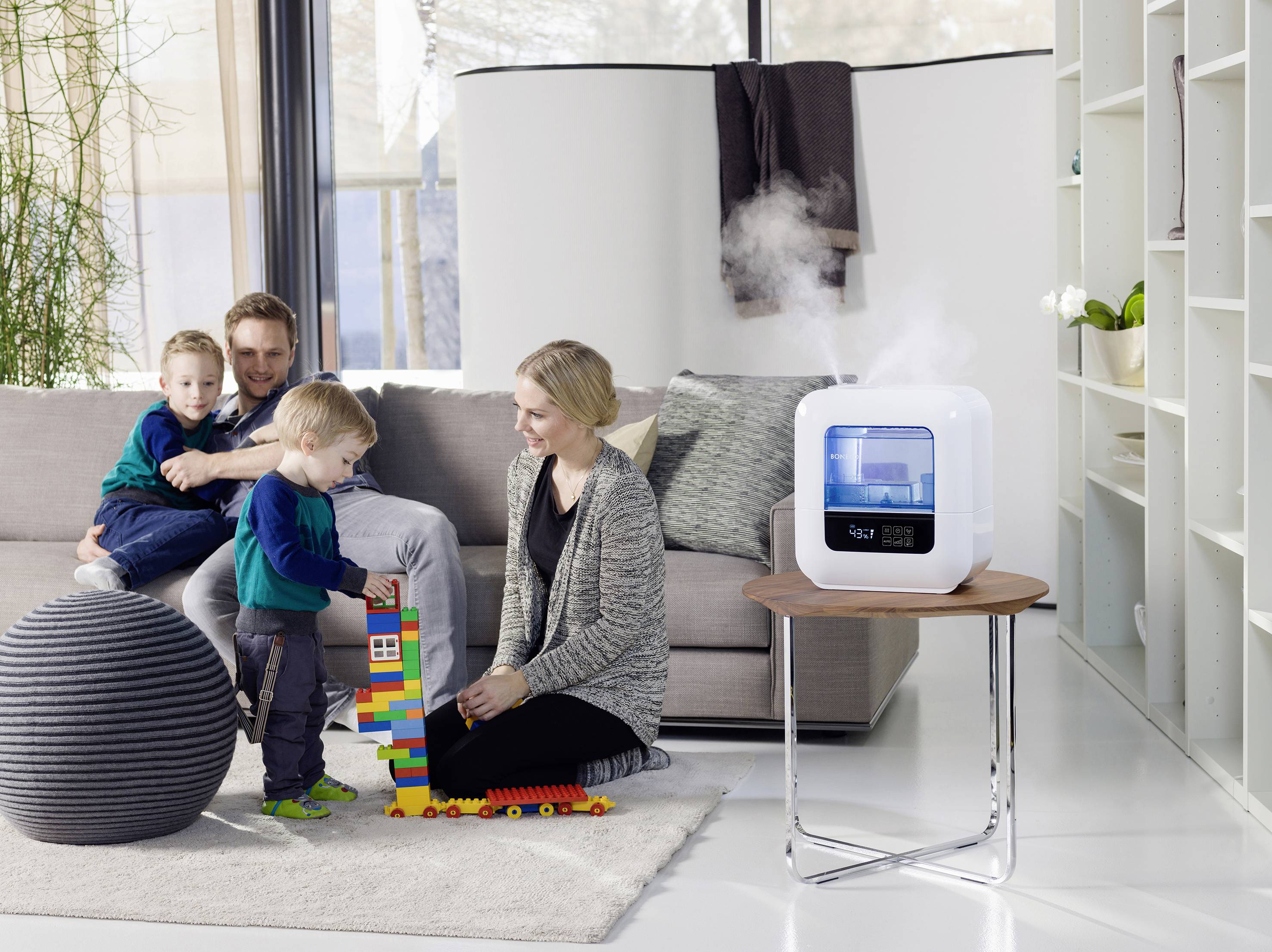 A family with two children is sitting in the living room. A humidifier is placed on a table and is emitting steam.