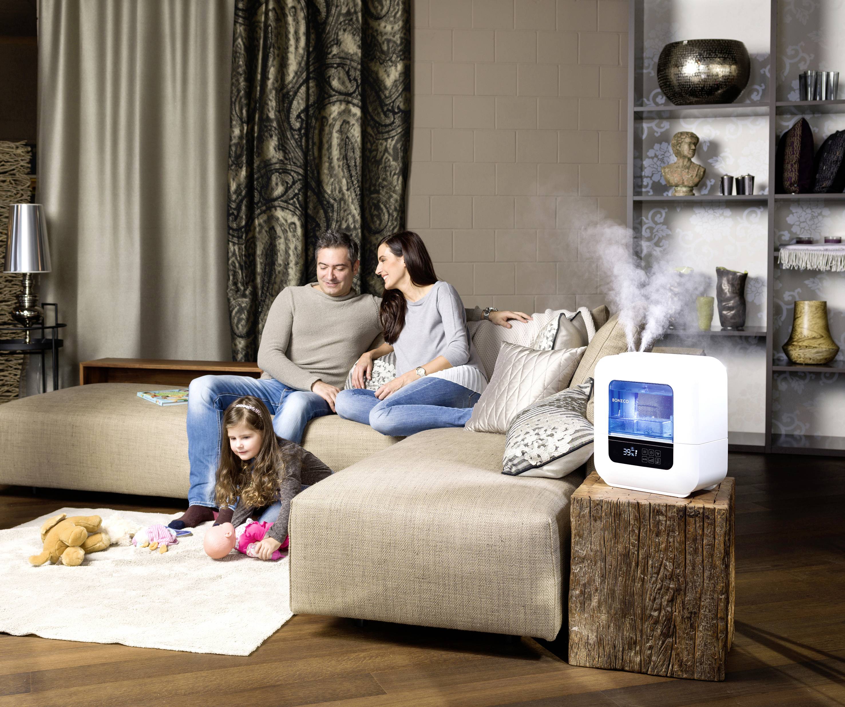 A family is sitting on a sofa in a living room. A humidifier is placed on a table nearby, with steam rising from it.