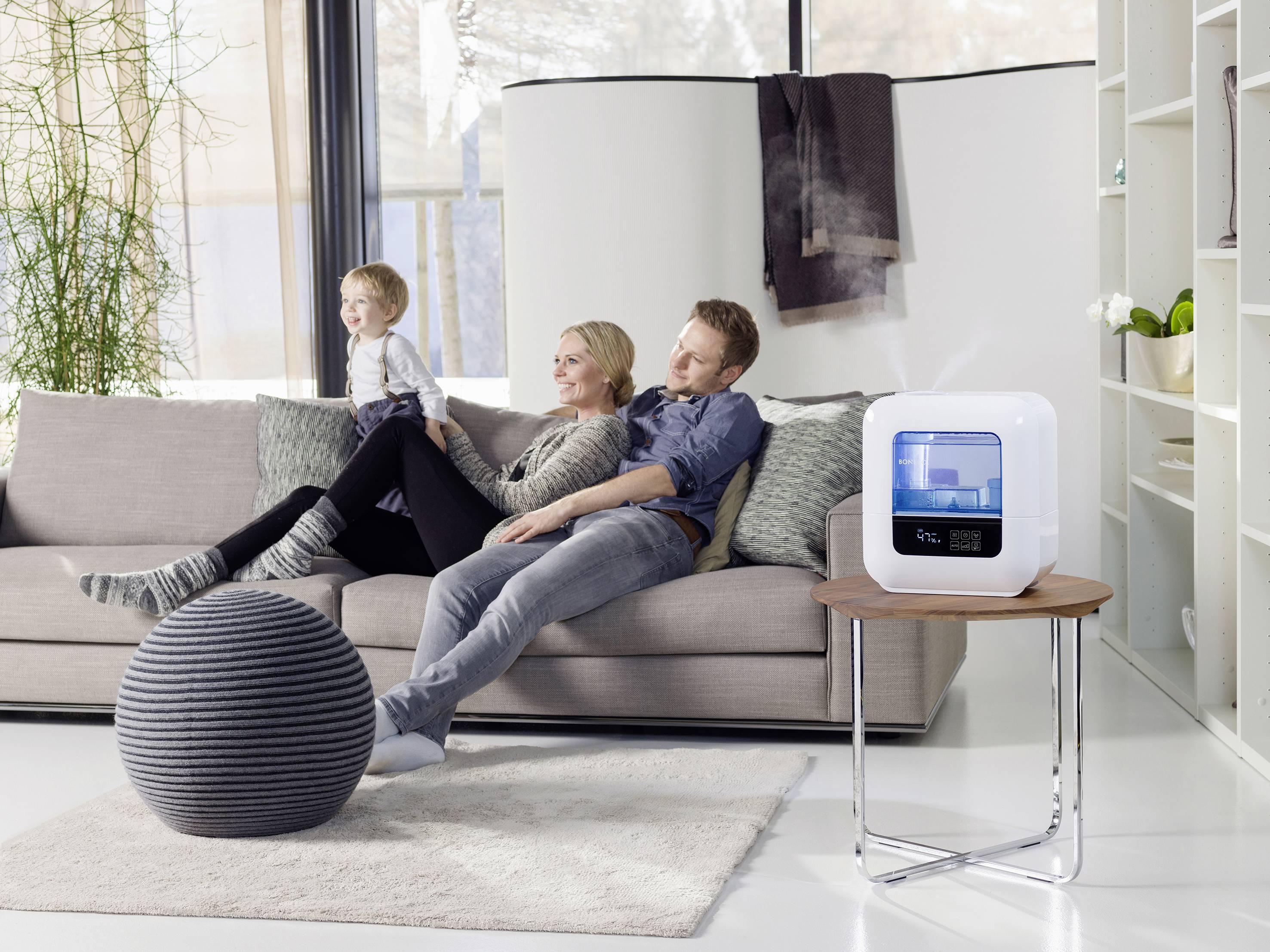 A family sits relaxed on a sofa in a modern living room. An air purifier stands on a table beside them.