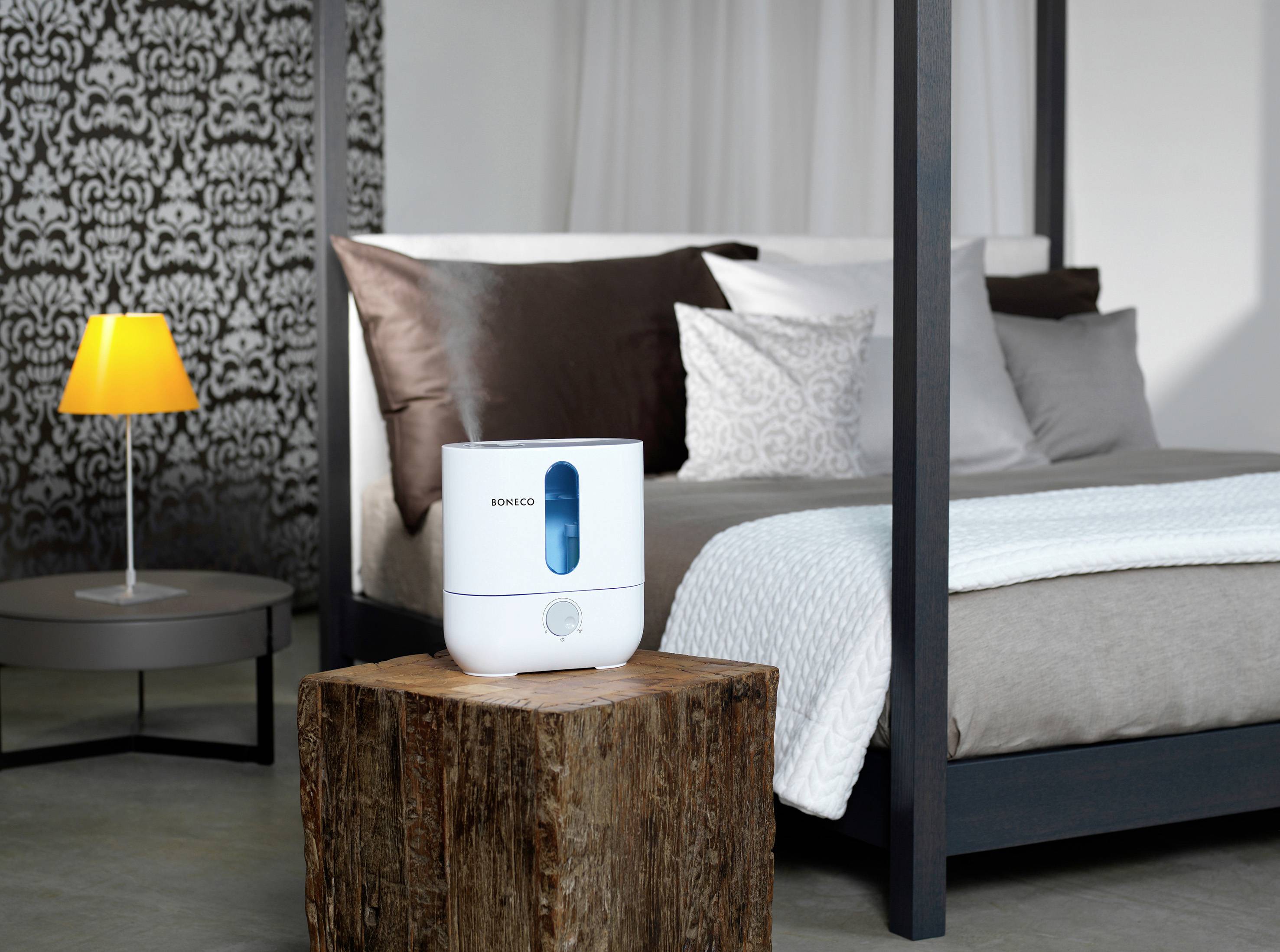 A humidifier sits on a wooden table in front of a bed in a modern-furnished bedroom. A lamp illuminates the background.
