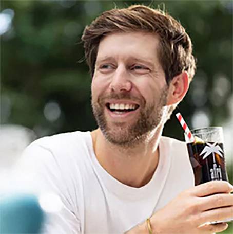 A person with a beard smiles while holding a glass with a dark drink and a straw. Background blurred.