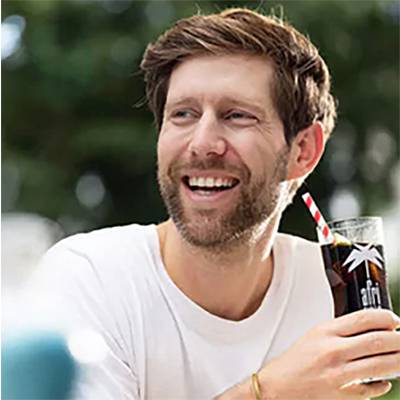 A person with a beard smiles while holding a glass with a dark drink and a straw. Background blurred.
