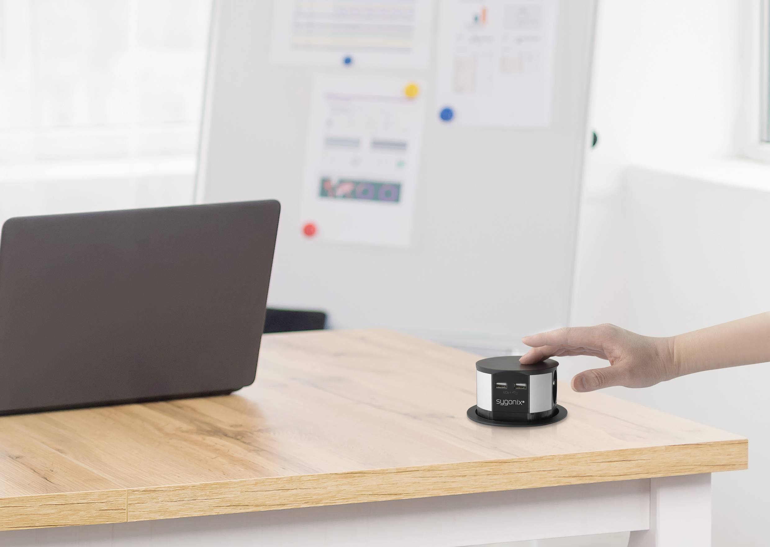 A person is pressing a grey button on a desk, next to a laptop. A whiteboard is in the background.