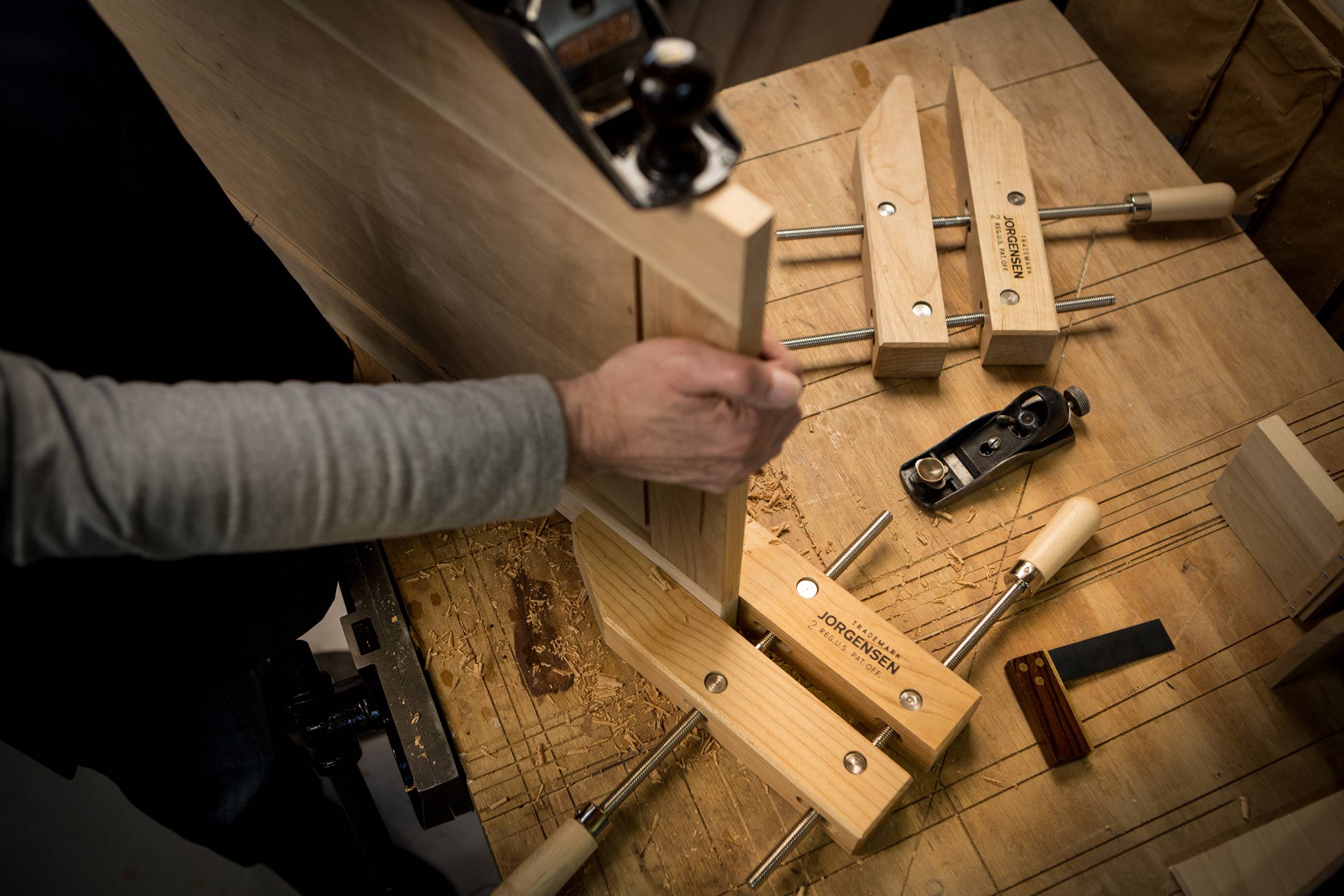 A person is working on a piece of wood in a workshop. Tools such as clamps and a plane can be seen on the workbench.
