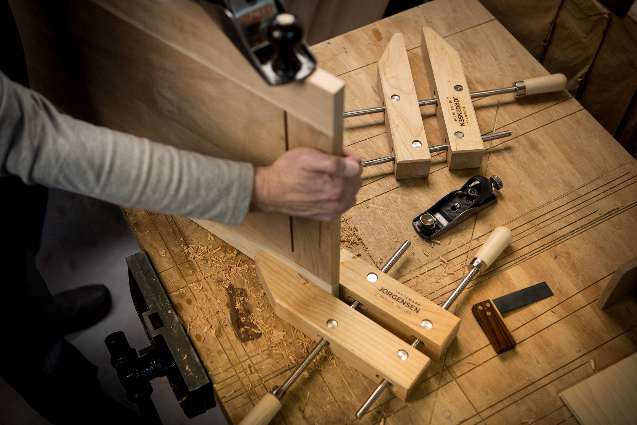 A person is working on a wooden piece on a workbench. Various tools, such as clamps and a plane, are scattered around.