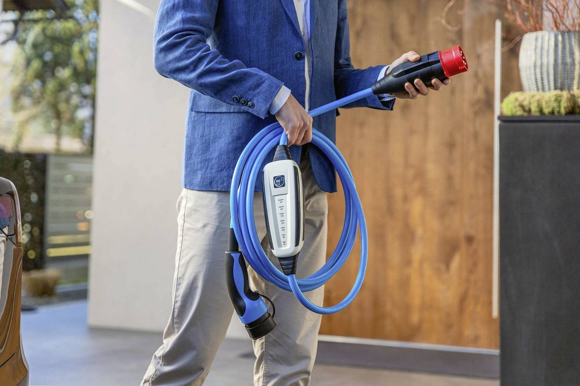 A person wearing a blue jacket is holding a blue charging cable for electric vehicles. In the background, there is wooden panelling and plants.