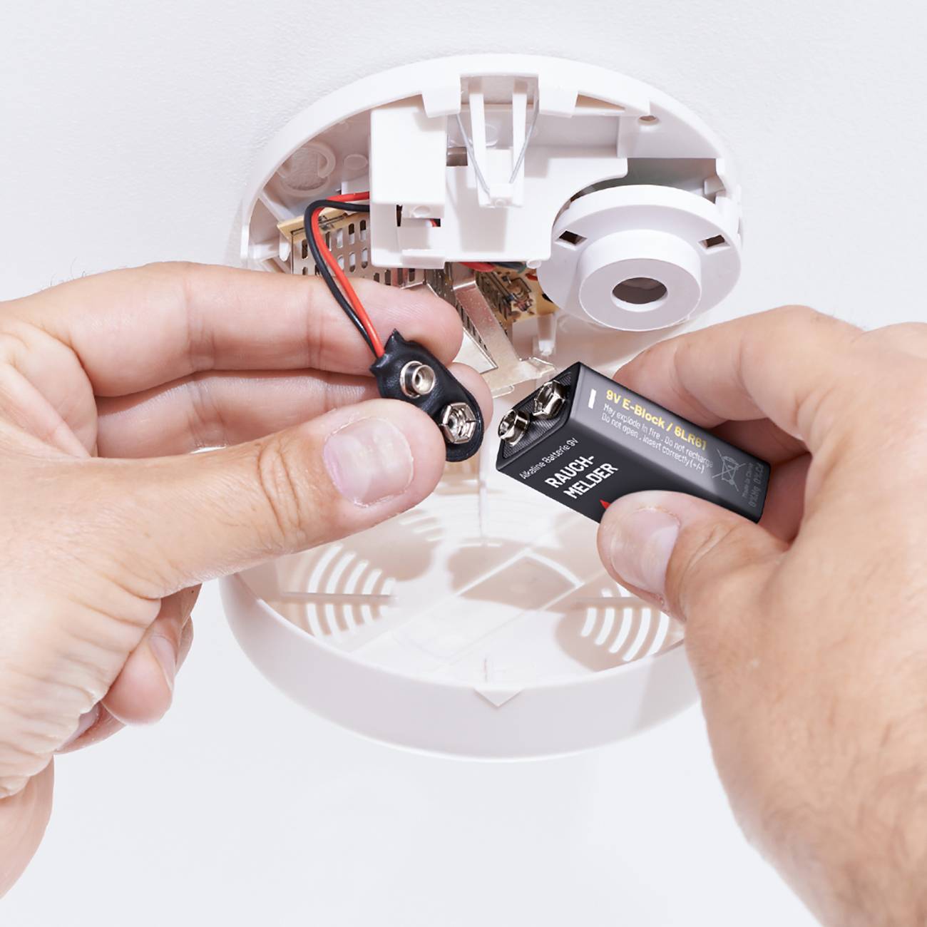 Two hands are installing a 9-volt battery in a white smoke detector on the ceiling.