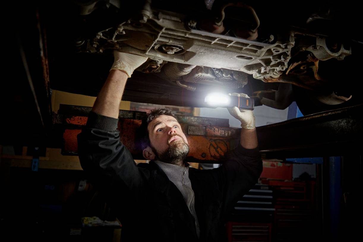 A mechanic is repairing a car underneath it in a garage. He is using a lamp to illuminate the area.
