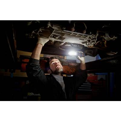 A mechanic is repairing a car underneath it in a garage. He is using a lamp to illuminate the area.