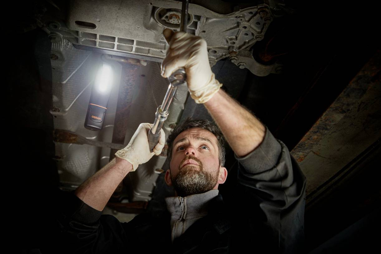 A mechanic is working underneath a car in the workshop, holding a spanner and wearing gloves.