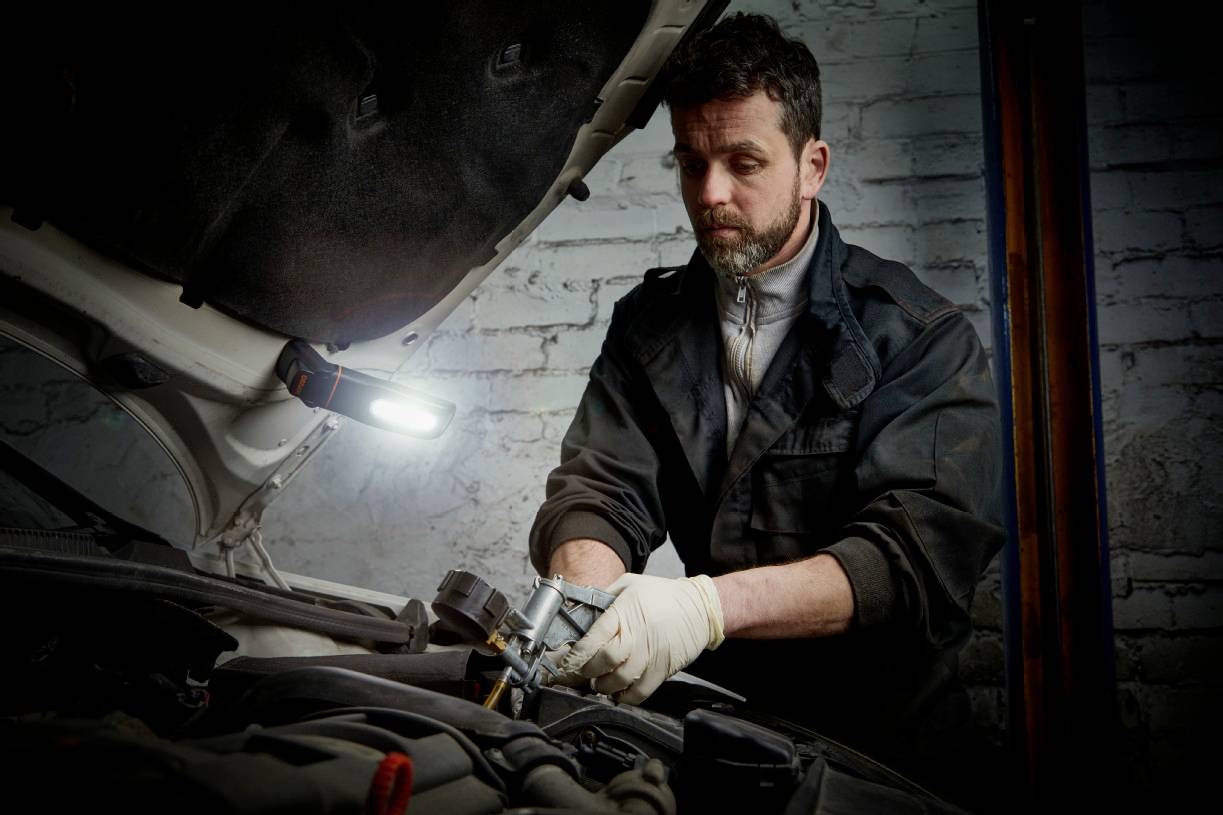 A mechanic in workwear is repairing an engine in a workshop, with a lamp illuminating the work area.