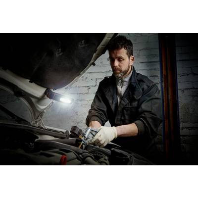 A mechanic in workwear is repairing an engine in a workshop, with a lamp illuminating the work area.