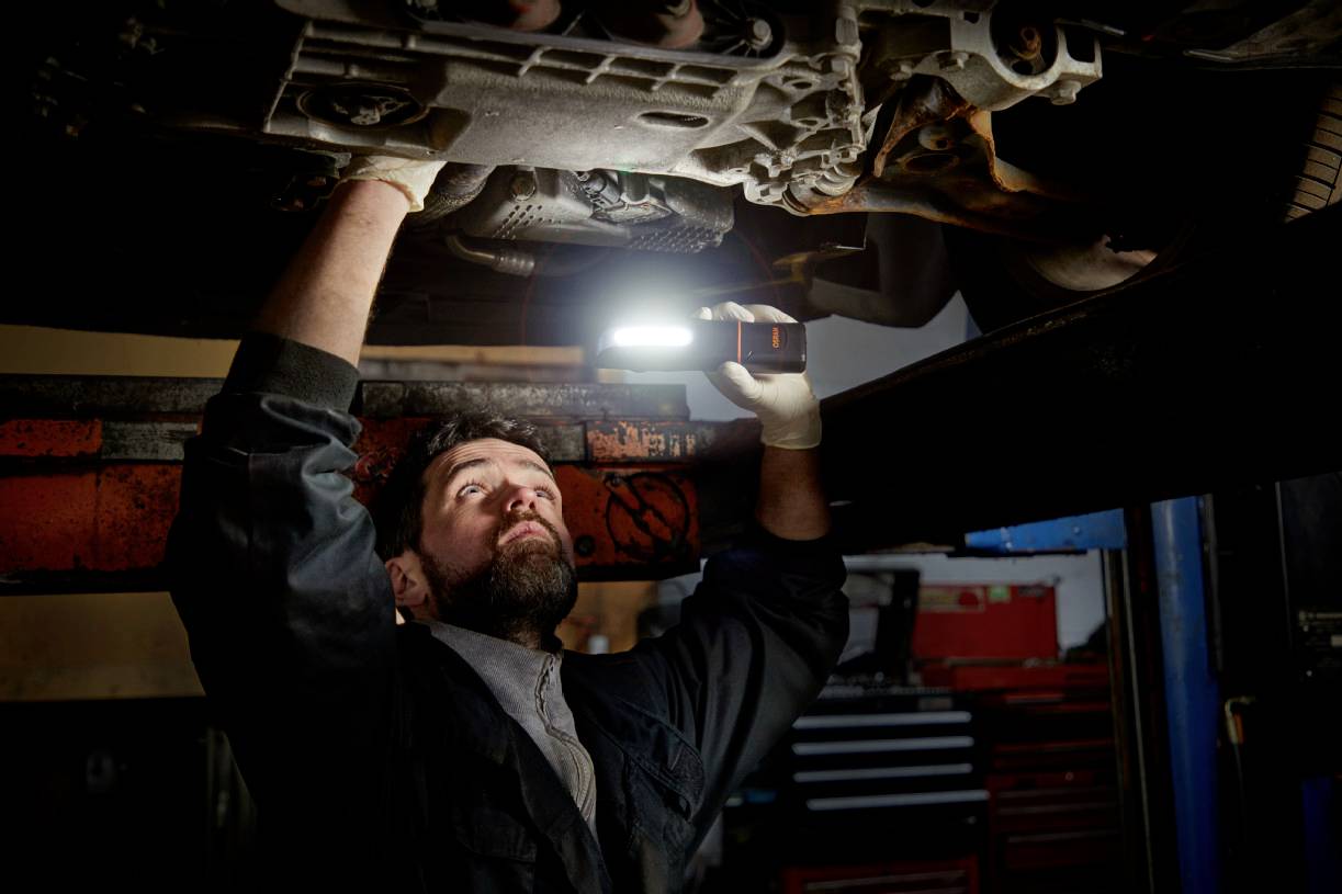 A mechanic inspects the underside of a vehicle in a workshop, holding a portable lamp.