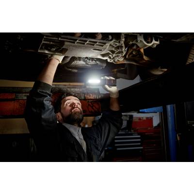 A mechanic inspects the underside of a vehicle in a workshop, holding a portable lamp.