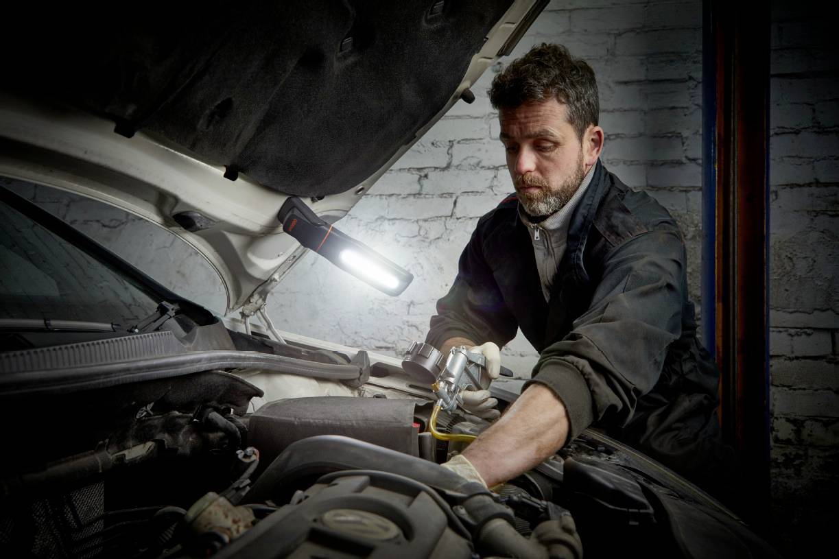A mechanic is examining the engine of a car in a workshop. A lamp is illuminating the engine compartment.