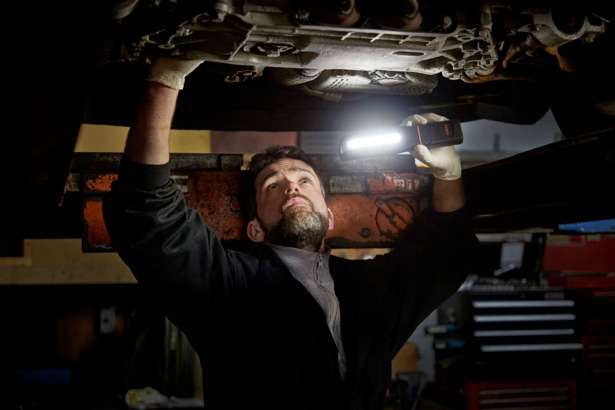 A mechanic is repairing underneath a vehicle and illuminating the area with a portable lamp in order to see better.