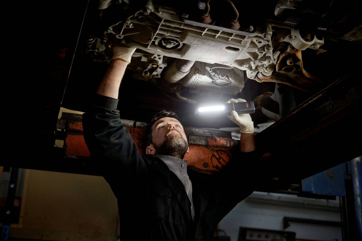 A mechanic is working underneath a vehicle and holding a torch to illuminate the engine compartment.