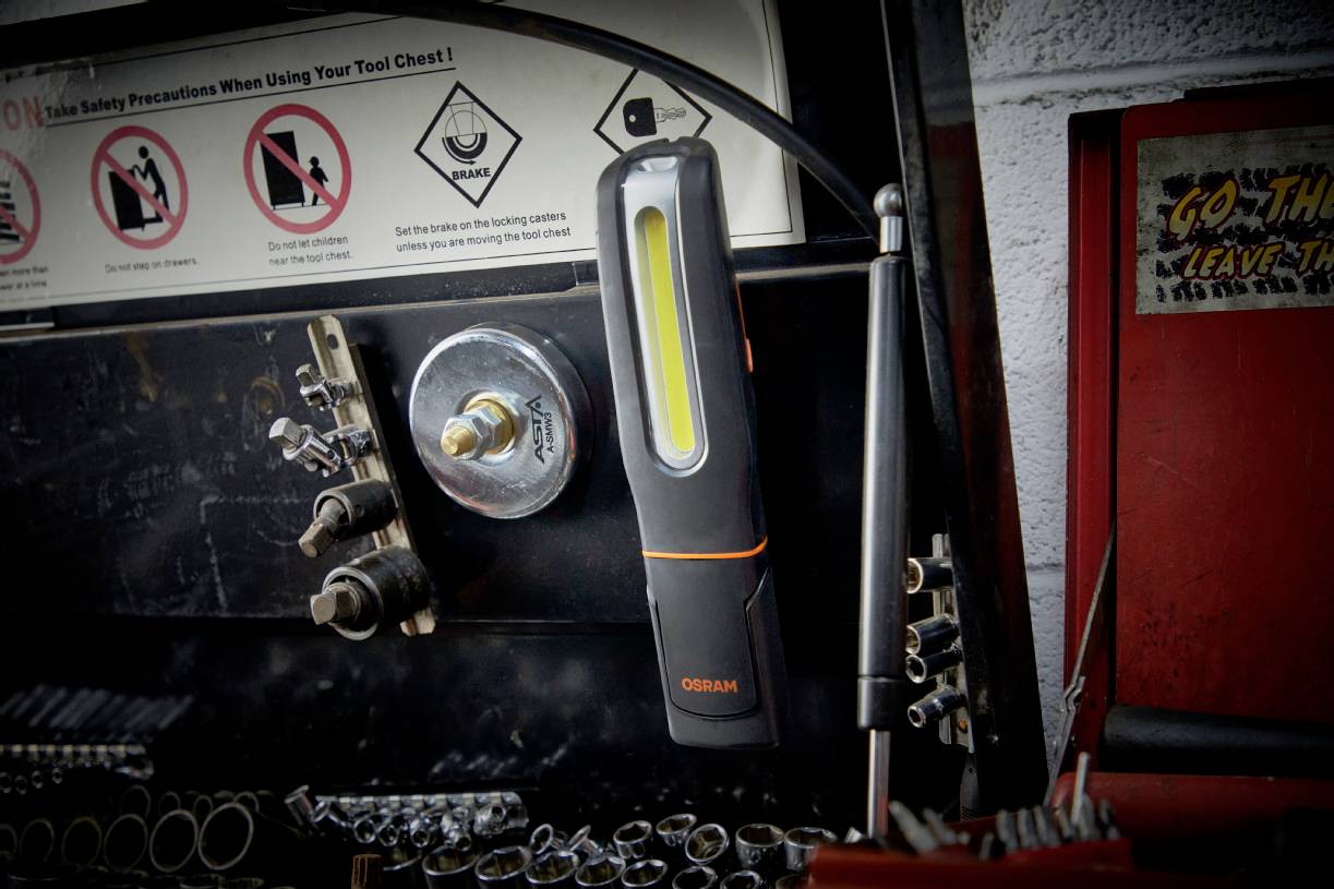 A portable work lamp hangs from a metallic surface in a workshop, surrounded by tools and safety instructions.