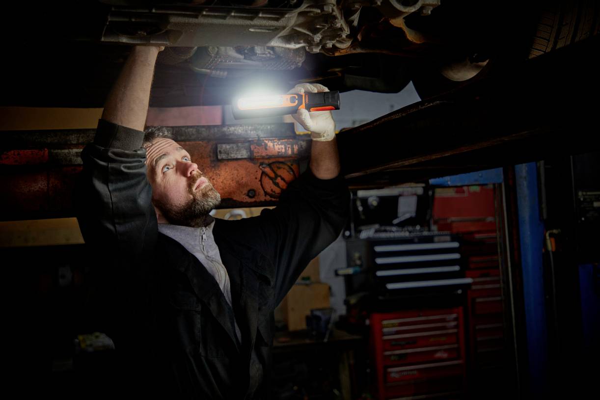 A mechanic is working in a workshop underneath a car, holding a lamp to illuminate the work area. Tools are visible in the background.